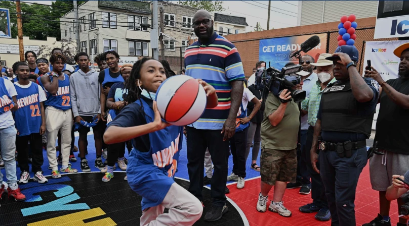 A young girl playing with a basket ball at one of the Newark Comebaq Court, while Shaquille O'Neil and several other people stand in the background.